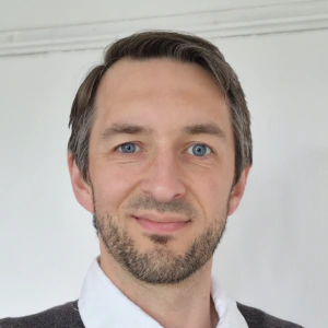 A man with short brown hair, blue eyes, and a trimmed beard wears a white collared shirt under a dark sweater, standing in front of a plain white background, smiling softly.