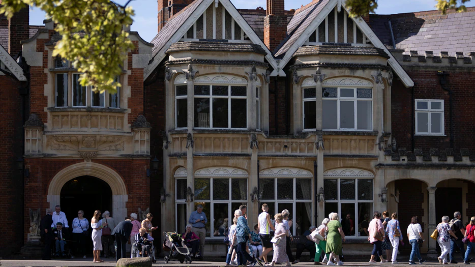 A group of people walk and gather outside a large historic brick building with ornate windows and gables on a sunny day, with leafy branches framing the scene.