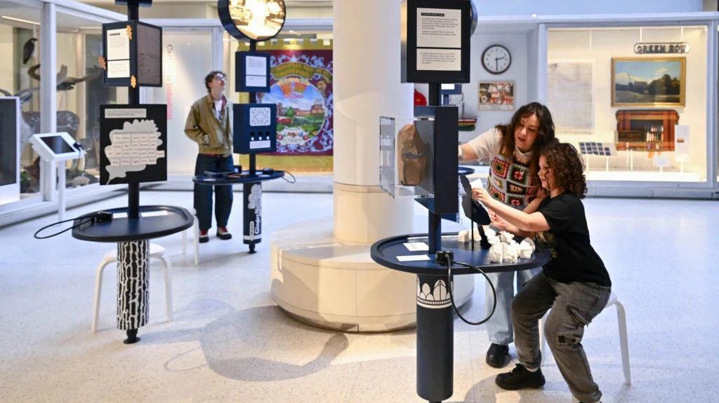 Three people interact with educational displays in a bright museum exhibit. Two people laugh together at one station while another person explores a display in the background. Colorful artwork decorates the walls.
