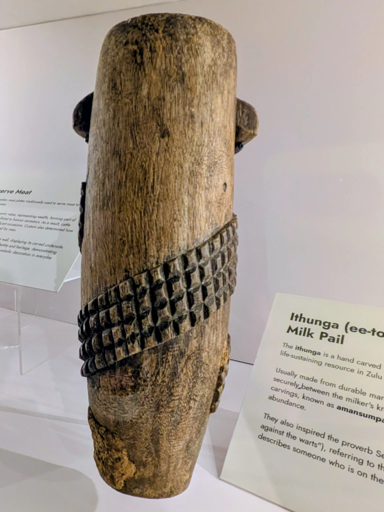 A carved wooden Zulu milk pail called Ithunga with a geometric patterned band, displayed in a museum with descriptive plaques in the background.
