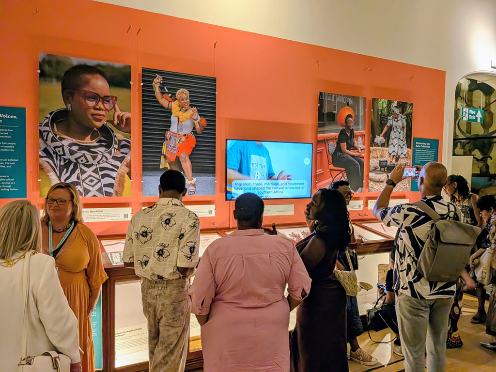 A group of people view an exhibit featuring three large portraits, a screen, and display cases with artifacts against an orange wall in a museum setting.