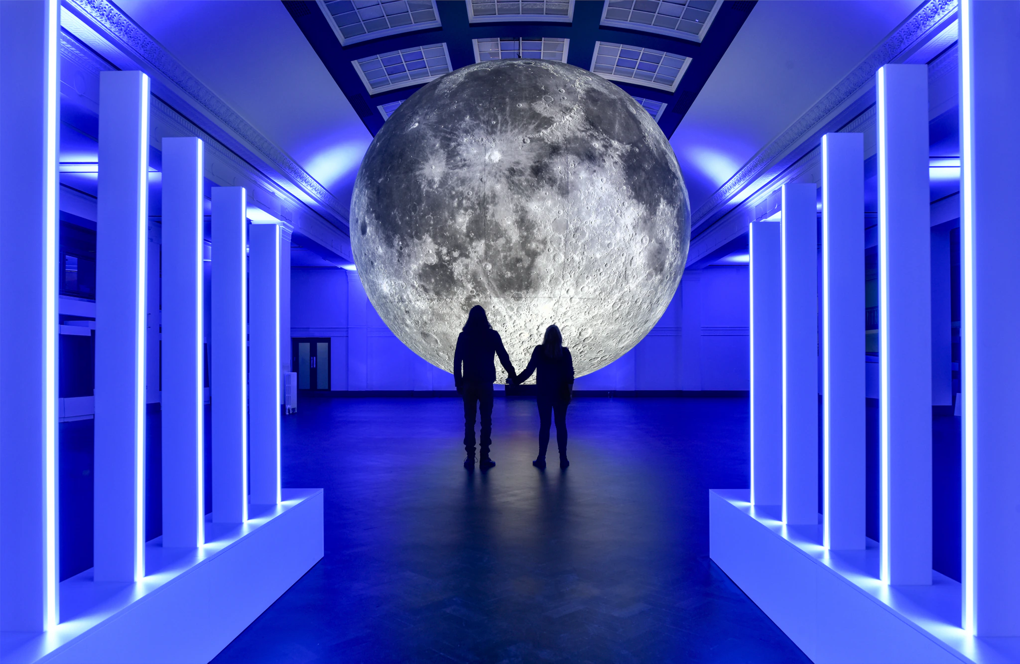 Two people look at a large moon sculpture in a blue-lit gallery space