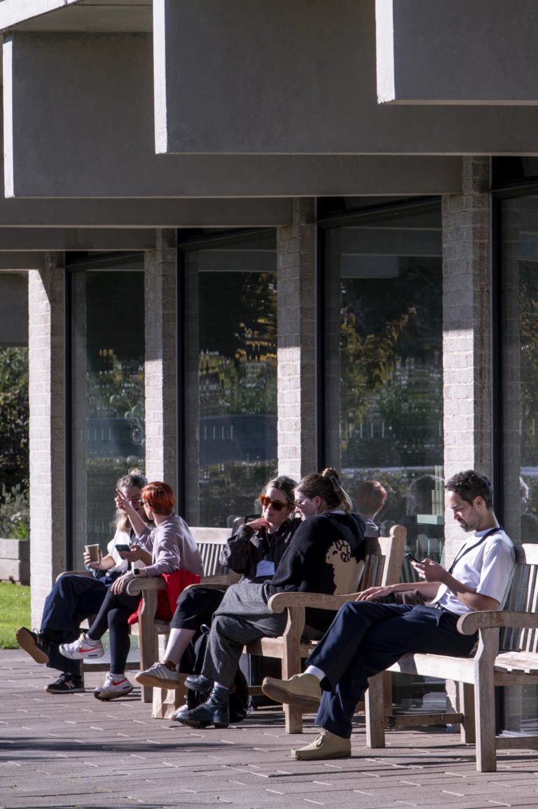 People relax on chairs outdoors, drinking coffee and using their phones, at an MA event